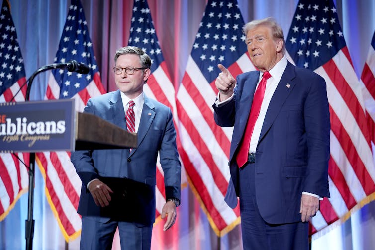 file-20241118-14-rrh5nu - The Deeping Two men in blue suits standing in front of many US flags.